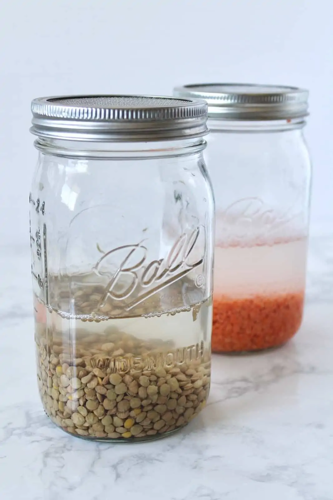 Two mason jars, one with red lentils and water, another with brown lentils and water, on a white marble surface