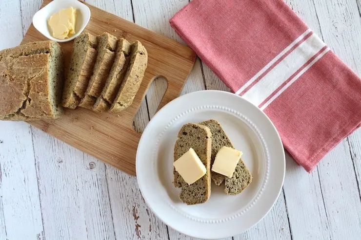 Slices of keto bread with slabs of butter on them on white plate next to loaf with slices out of it on wooden cutting board
