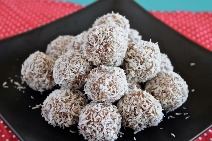 Close up of a pile of coconut date balls on a black plate on top of a red mat with white polka dots on a blue table