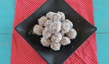 A pile of coconut date balls on a black plate on top of a red mat with white polka dots on a blue table