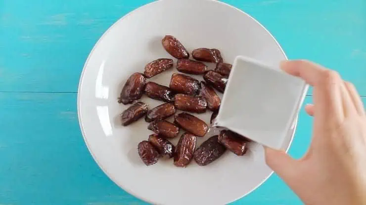 A hand pouring water over dates in a white bowl on a blue table