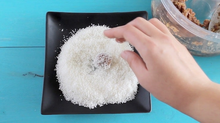Black plate full of shredded coconut on a blue table with a hand rolling a coconut date roll on the pile of coconut