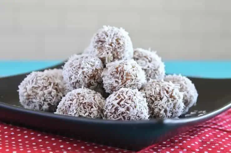 Close up of a pile of coconut date balls on a black plate on top of a red mat with white polka dots on a blue table with a white subway tile background