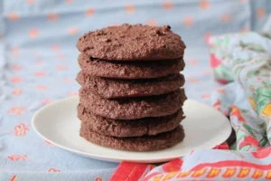 Six gluten free chocolate cookies stacked on top of each other on a white plate surrounded by blue table cloth with a red floral border