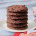 Six gluten free chocolate cookies stacked on top of each other on a white plate surrounded by blue table cloth with a red floral border