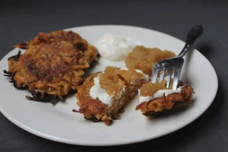 Close up of latke on a white plate with sour cream and applesauce on top of it with a fork in it