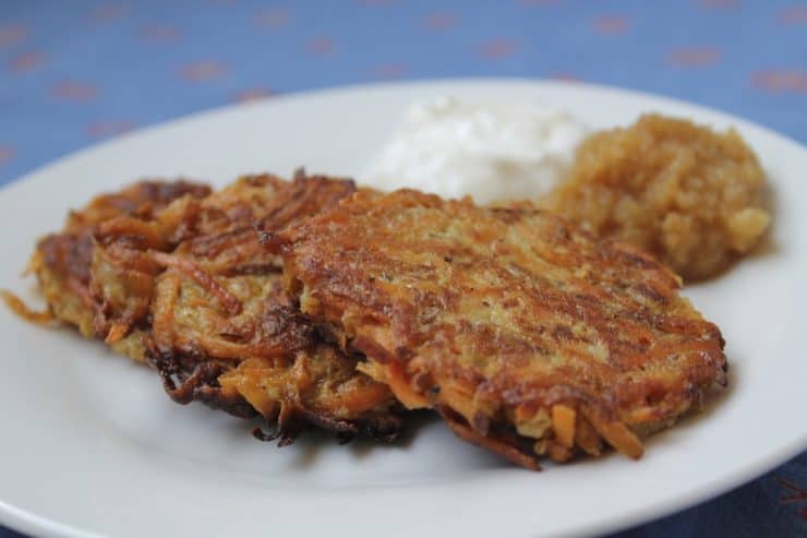 closeup of white plate with 3 latkes on it with a dollop of sour cream and applesauce over a blue tablecloth