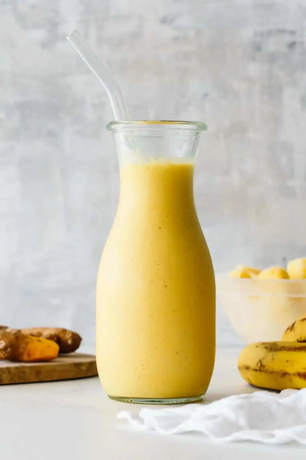 carafe filled with orange colored turmeric pineapple smoothie on a white table next to bananas and turmeric root