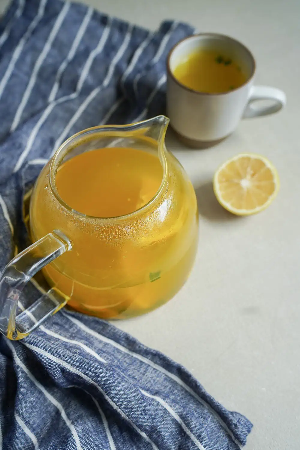 pitcher of yellow turmeric tea with lemon slices in it on a white surface next to a halved lemon and a blue and white dish towel