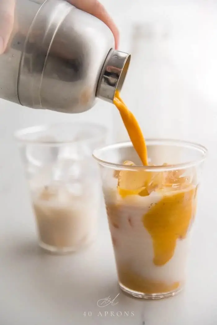 stainless steel tumbler pouring turmeric drink into a clear glass with milk inside 