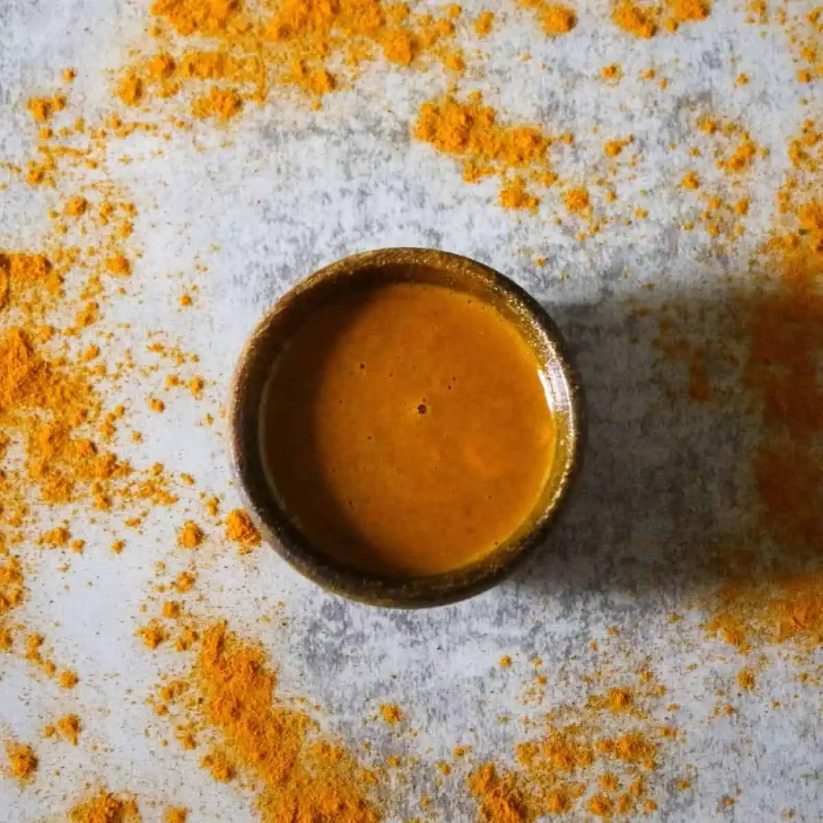 overhead shot of small bowl with orange colored turmeric mask liquid inside on a grey surface with sprinkled turmeric around it