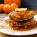 A stack of paleo pumpkin pancakes on a plate with maple syrup and pumpkins in the background