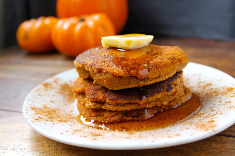 A stack of paleo pumpkin pancakes on a plate with maple syrup and pumpkins in the background