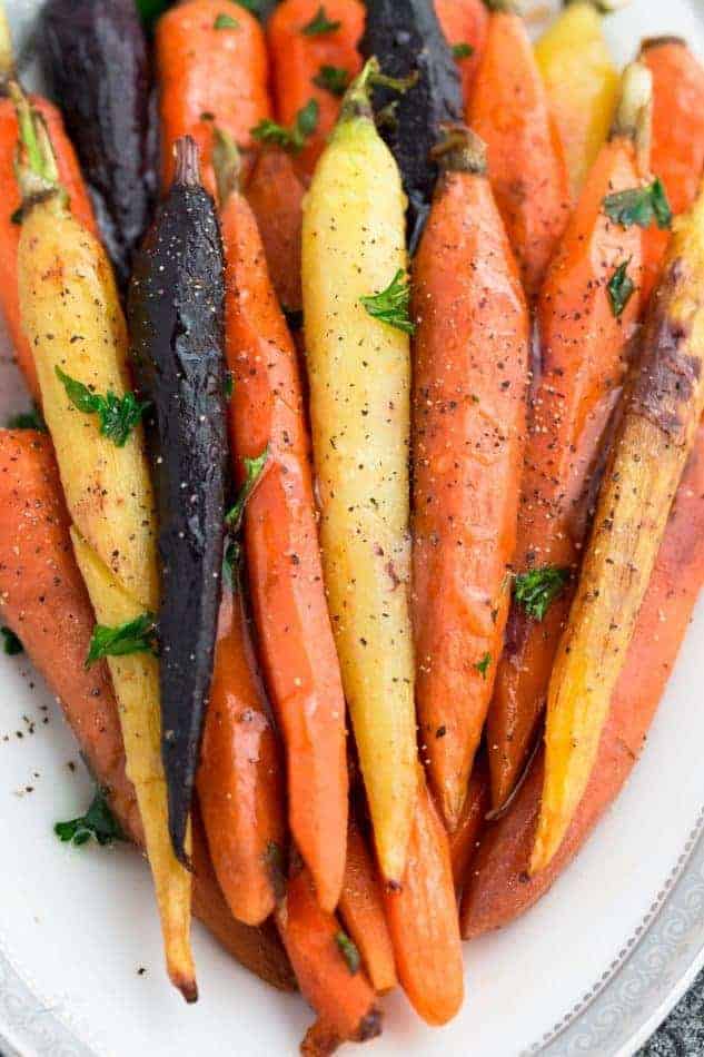 Whole roasted different colored carrots on a white plate arranged in the same direction on top of eachother with spices and herbs sprinkled on top 