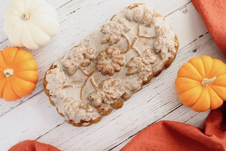 Overhead view of whole loaf of healthy paleo pumpkin bread with white icing on top on white wooden surface next to small pumpkins and red dish towel