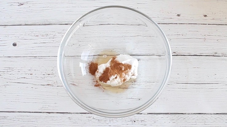 Clear glass bowl on a white wooden surface with coconut cream, cinnamon and syrup inside it