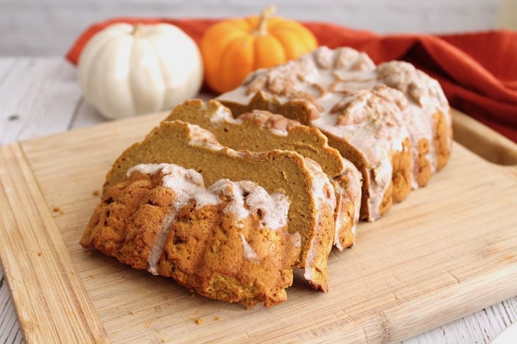 Sliced loaf of healthy paleo pumpkin bread with white icing on top on wooden cutting board with small pumpkins in the background