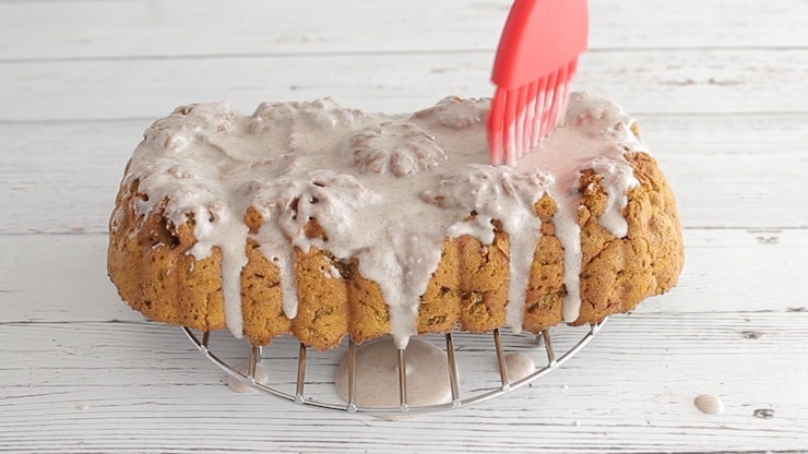 Red brush spreading white icing over paleo pumpkin bread sitting on metal wire over a white wooden surface