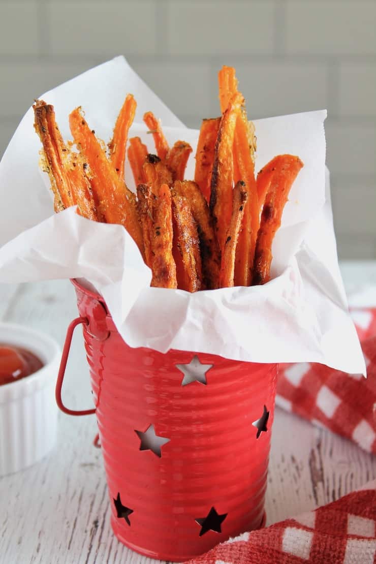 Carrot fries in a small red bucket lined with parchment paper next to white ramekin of ketchup on white table