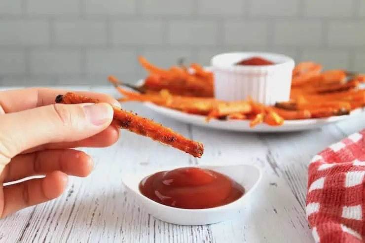 Hand holding a carrot fry about to dip in a ramekin of ketchup with a plate of carrot fries in the background