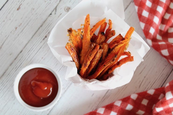 overhead view of Carrot fries in a small red bucket lined with parchment paper next to white ramekin of ketchup and red and white checkered napkin on white table