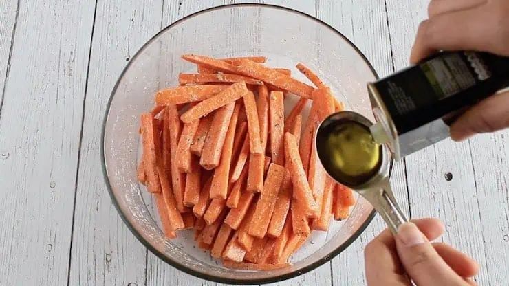 sliced carrots with spices mixed in in a clear bowl with a hand holding a tablespoon of olive oil over it