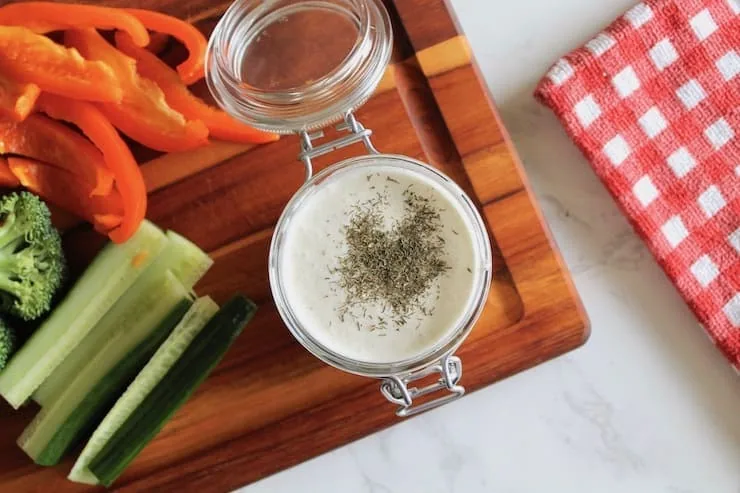 overhead shot of paleo ranch dressing in a jar on a wooden cutting board with sliced vegetables next to it