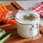close up of paleo ranch dressing in a jar on a wooden cutting board with sliced vegetables next to it