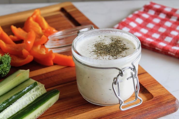 close up of paleo ranch dressing in a jar on a wooden cutting board with sliced vegetables next to it