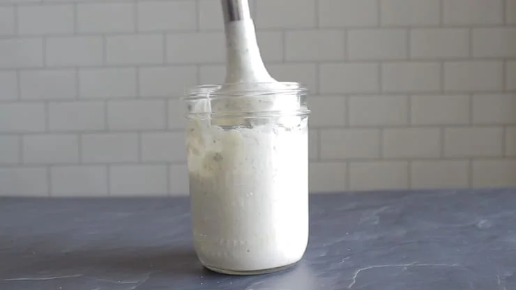 Glass jar filled with paleo ranch ingredients with hand blender in jar on black stone with tile background
