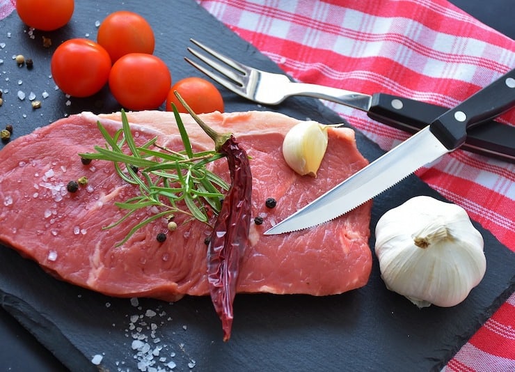 Raw seak with rosemary, chili pepper and garlic on it on a black slate with a red and white checkered napkin