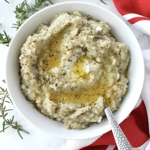 A bowl of cauliflower puree with a spoon and sprigs of rosemary.