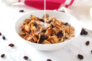 White bowl filled with aip raisin bran breakfast cereal with milk pouring on top of it on a white marble surface with raisin sprinkled on it next to a pink and white dish towel