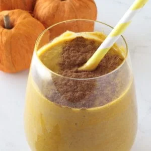 Close up of glass with orange pumpkin smoothie inside with cinnamon sprinkled on top and yellow and white striped straw inside