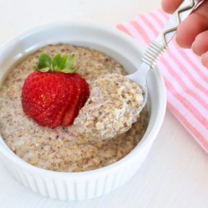 Spoon in bowl of low carb oatmeal in white bowl with strawberry on top next to pink striped towel