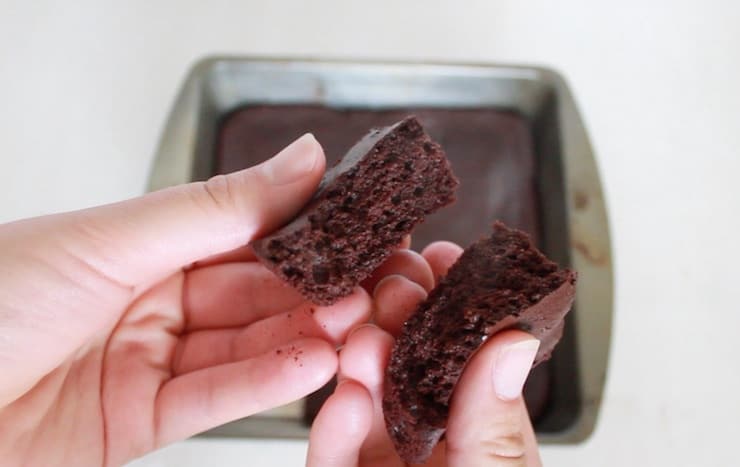 Close up of hands holding two halves of a freshly baked brownie with the pan in the background