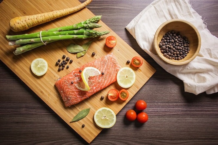 an overhead shot of a salmon fillet on a wooden board topped with a slice of lemon and vegetables around it