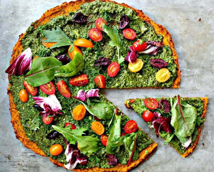 An overhead shot of a round healthy pizza crust topped with a green sauce, tomatoes and other vegetables