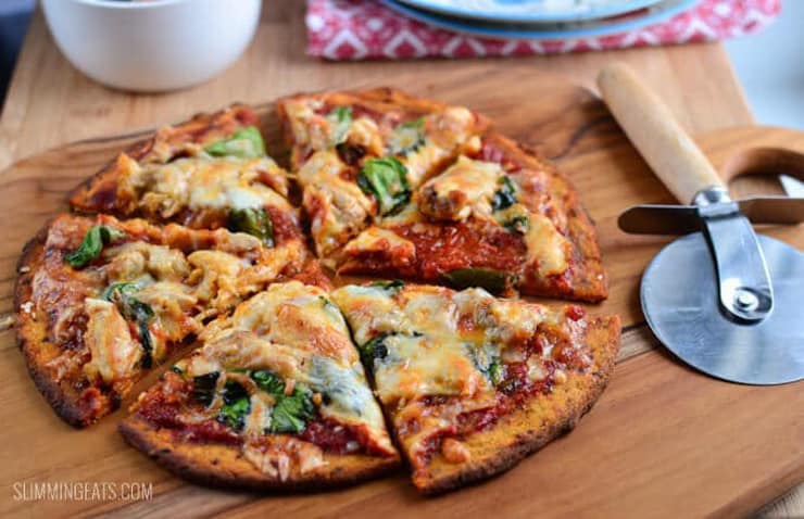 A round sweet potato healthy pizza crust cit into slices on a wooden board with a pizza cutter at the side