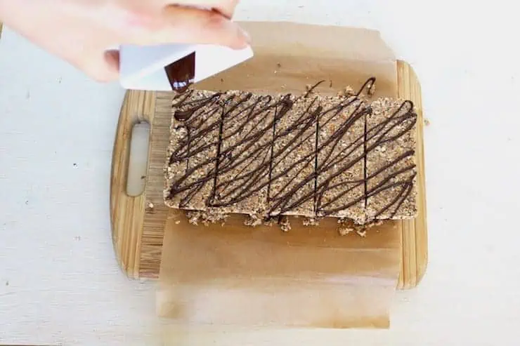 Sliced slab of homemade paleo protein bars being drizzled with chocolate on parchment paper on a wooden cutting board on a white wooden surface