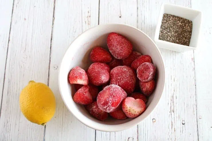 White bowl of frozen strawberries next to small ramekin of chia seeds and a lemon on a white wooden table