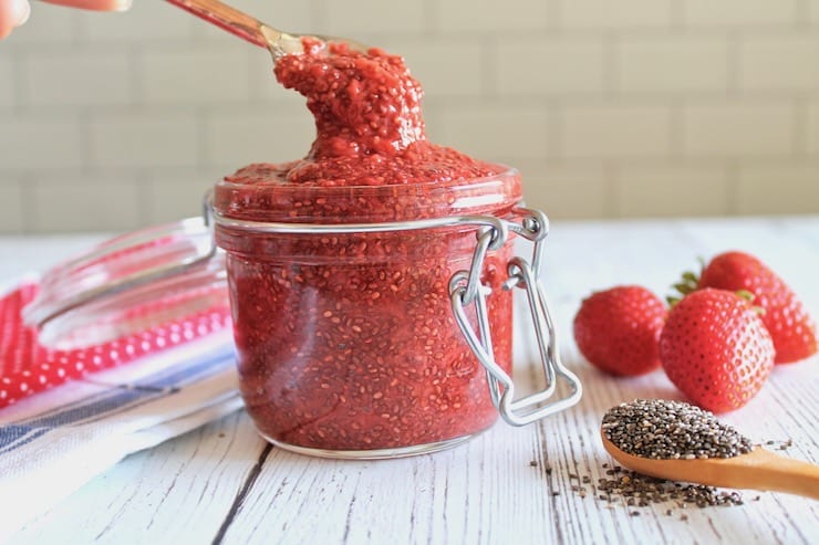 Spoon dipping into jar filled with chia strawberry jam on white wooden table next to fresh strawberries and wooden spoon of chia seeds