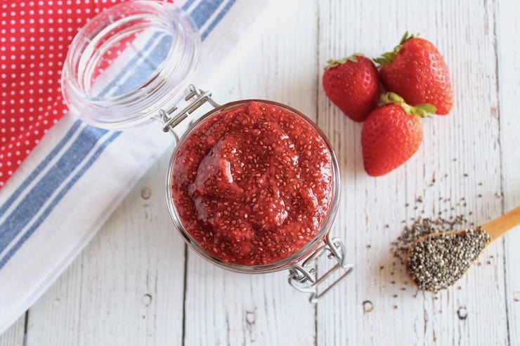 over head view of Jar filled with chia strawberry jam on white wooden table next to fresh strawberries and wooden spoon of chia seeds