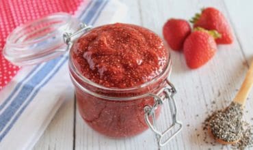 Jar filled with chia strawberry jam on white wooden table next to fresh strawberries and wooden spoon of chia seeds