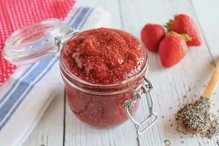 Jar filled with chia strawberry jam on white wooden table next to fresh strawberries and wooden spoon of chia seeds