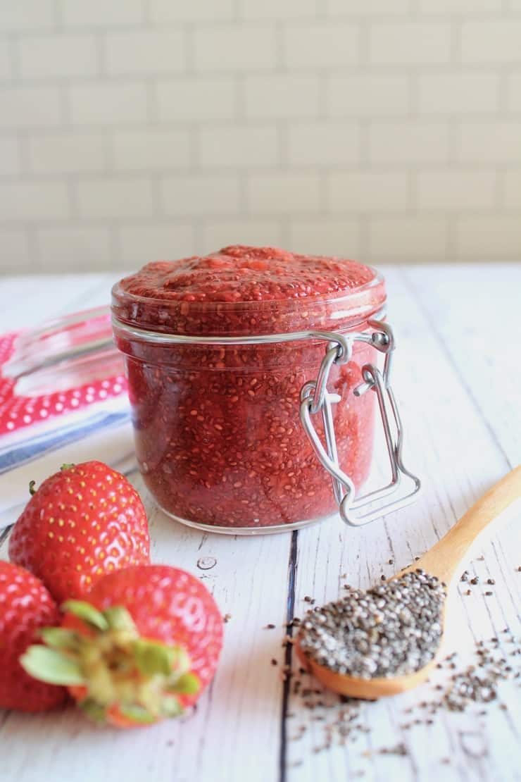 Jar filled with chia strawberry jam on white wooden table next to fresh strawberries and wooden spoon of chia seeds