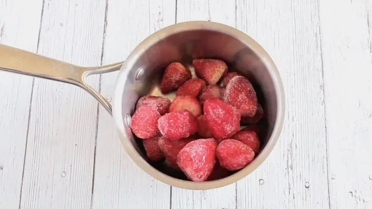 Stainless steel pot filled with frozen strawberries on a white wooden surface