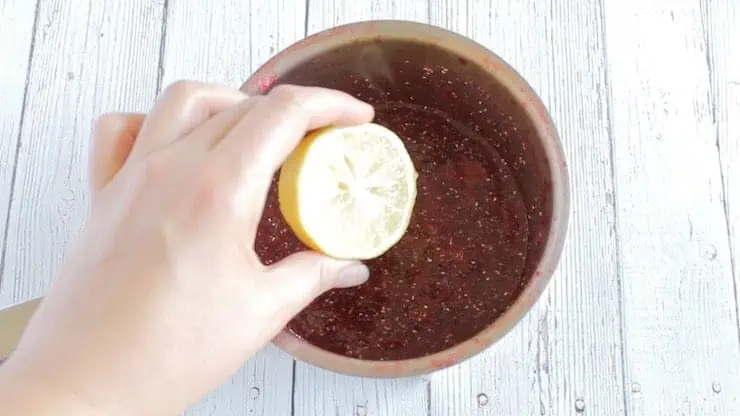 Hand holding half a lemon over stainless steel pot with chia strawberry jam ingredients inside