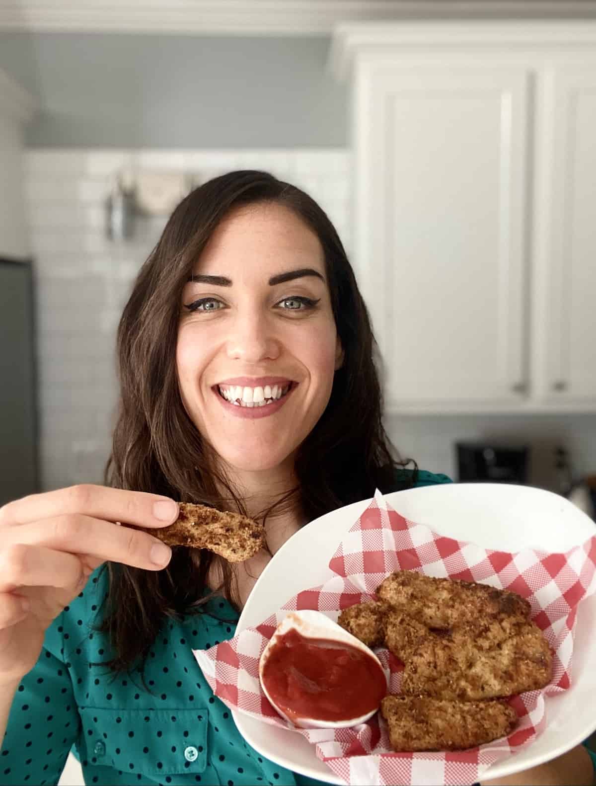 Smiling woman holding plate of chicken tenders and holding one tender with the other hand