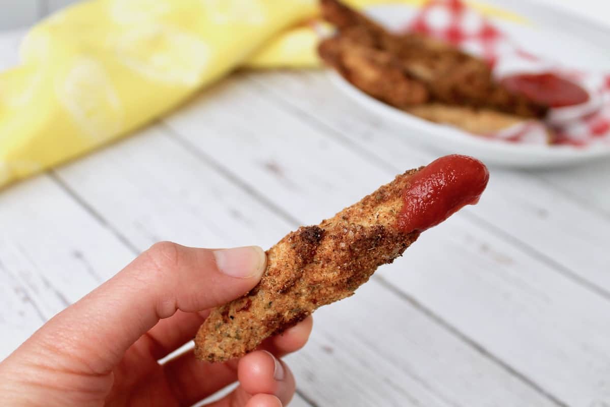 Close up of hand holding a chicken tender dipped in ketchup over a white wooden surface with a bowl of chicken tenders and a yellow dish towel in the background